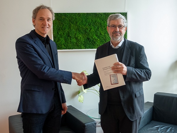 Prof. Dr. Bernd Hirschl übergibt an Minister Axel Vogel das Gutachten zum Klimaplan Brandenburg. Beide geben sich die Hand und lächeln in die Kamera.